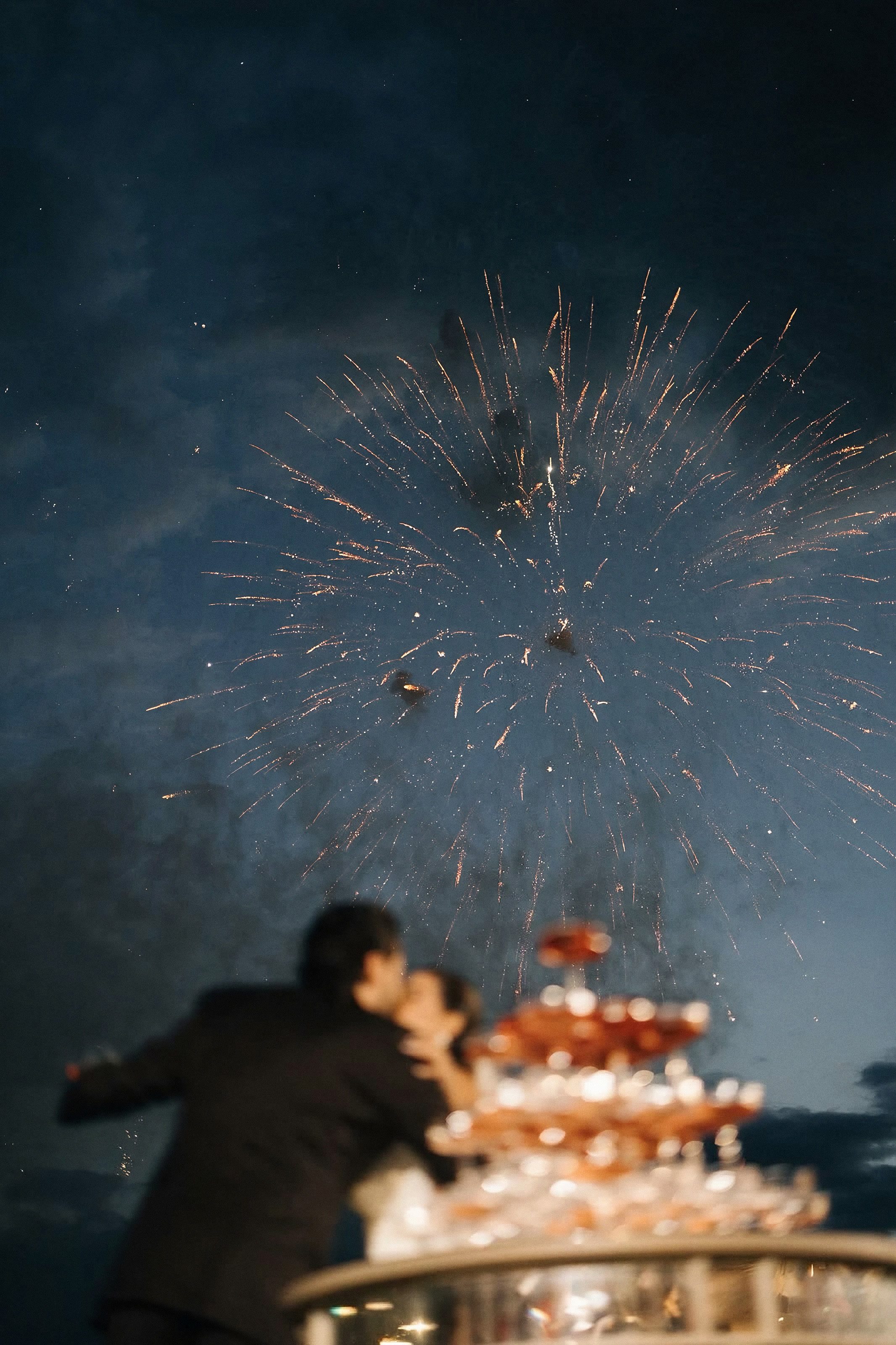 Noivos se beijando ao lado da torre de taças, com fogos de artifício no céu