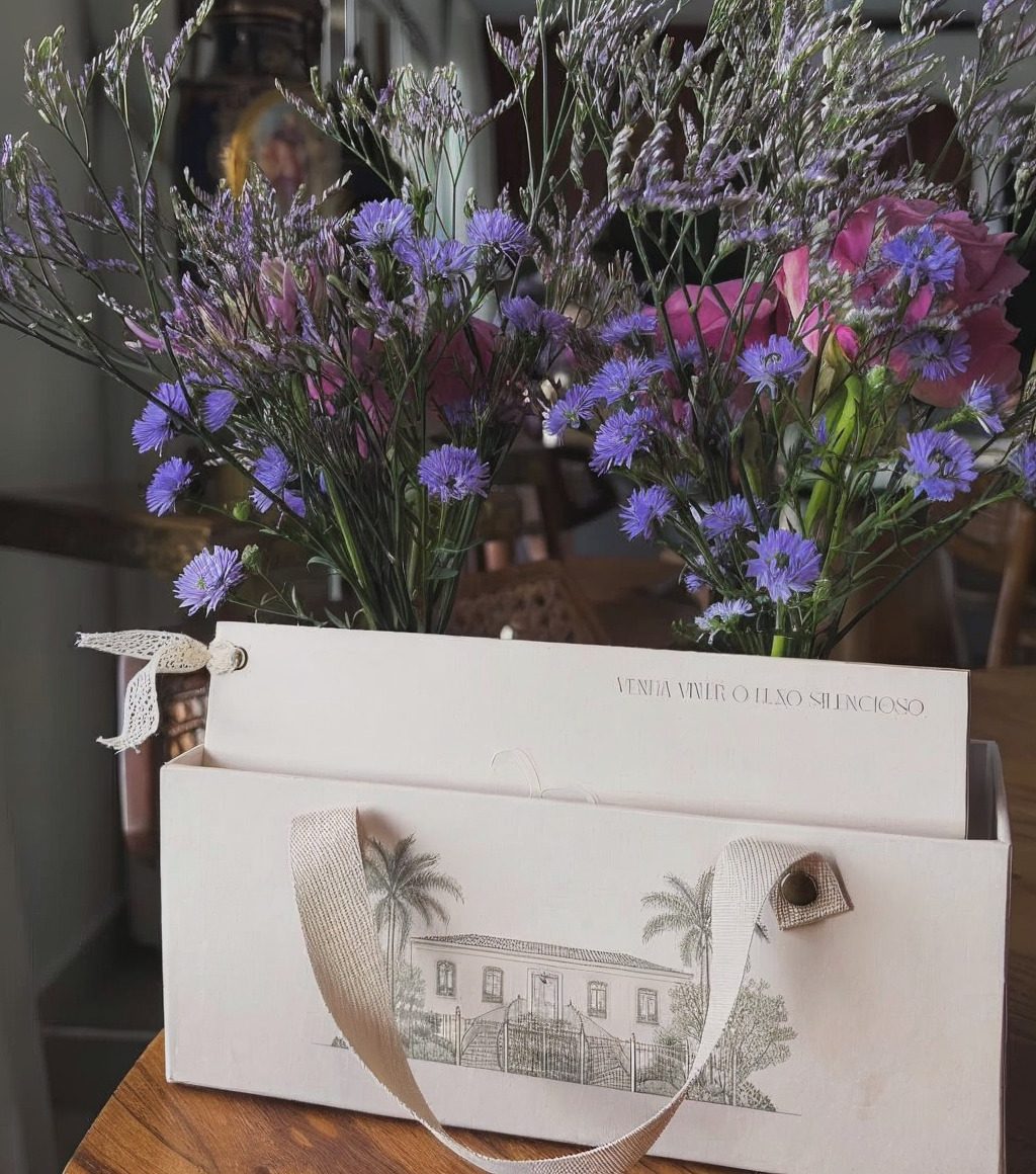 Caixa-convite da Versat Celebration com ilustração de casarão, fita e flores lilás, com a frase "Venha viver o luxo silencioso"