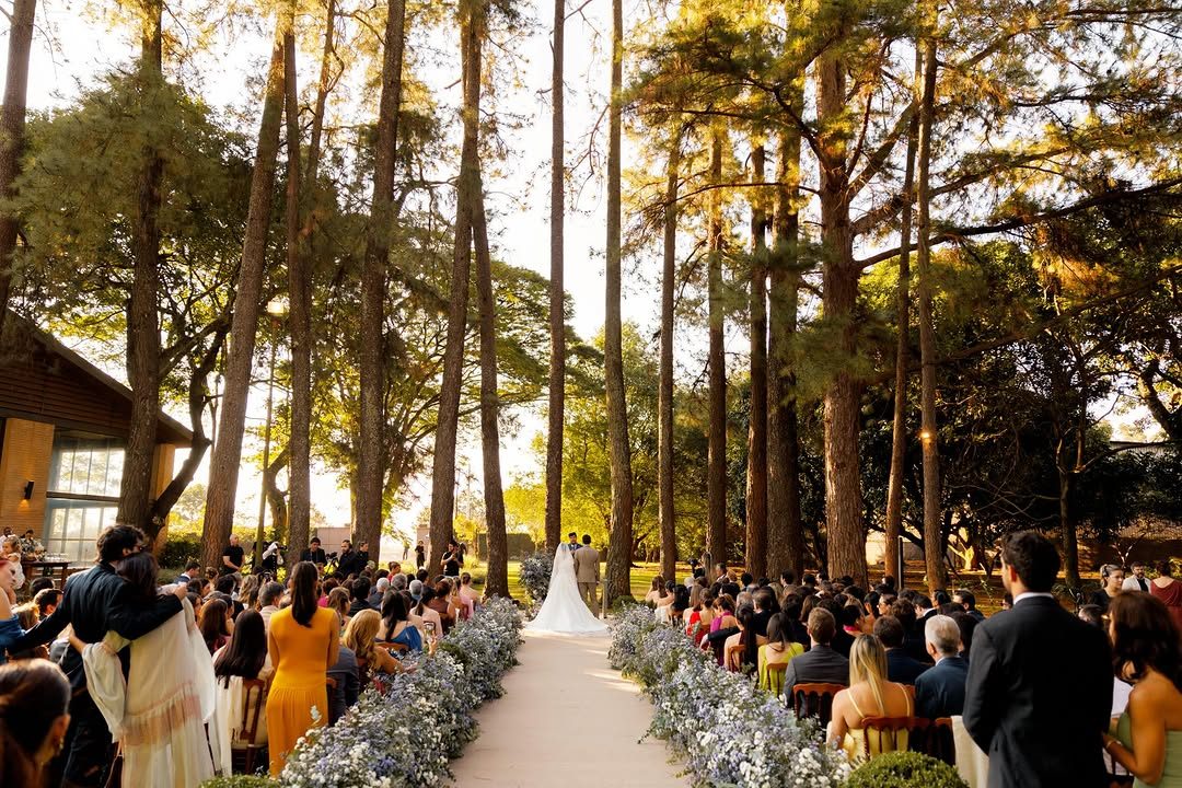 Cerimônia ao ar livre entre árvores altas, com convidados dos dois lados do corredor de flores