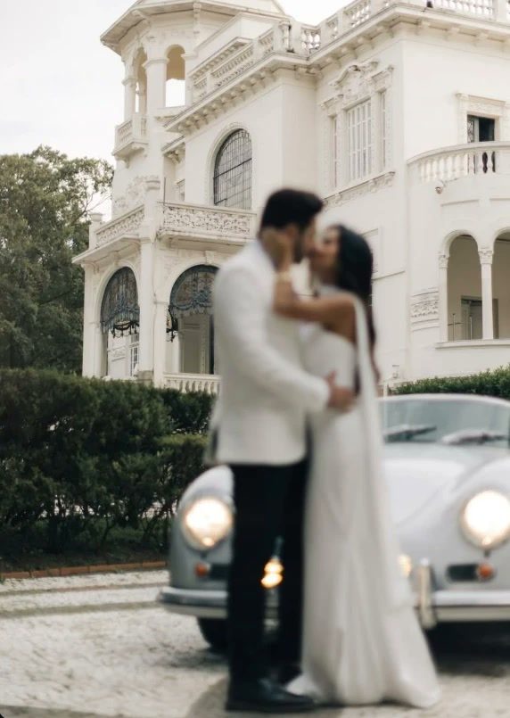 Noivos se beijando diante de um palacete branco e de um carro antigo