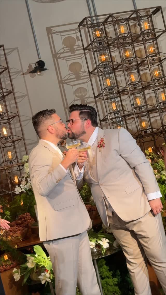 Dois noivos de terno claro brindando e se beijando
